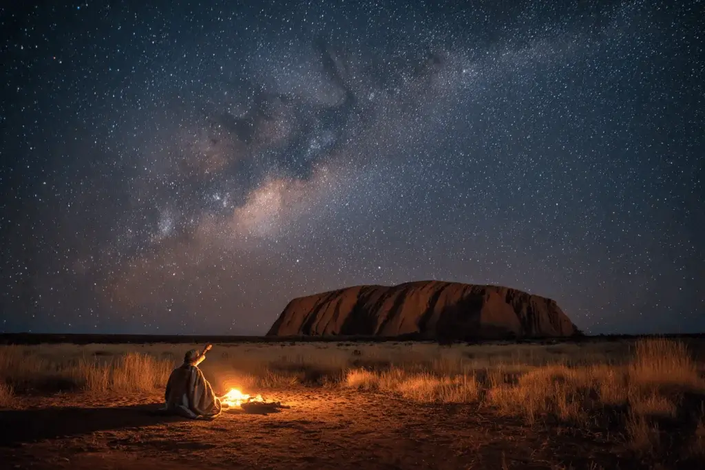 An Anangu starguide points toward the Emu in the Sky constellation above Uluru, the dark dust lanes of the Milky Way forming the emu’s shape in a clear desert night sky.